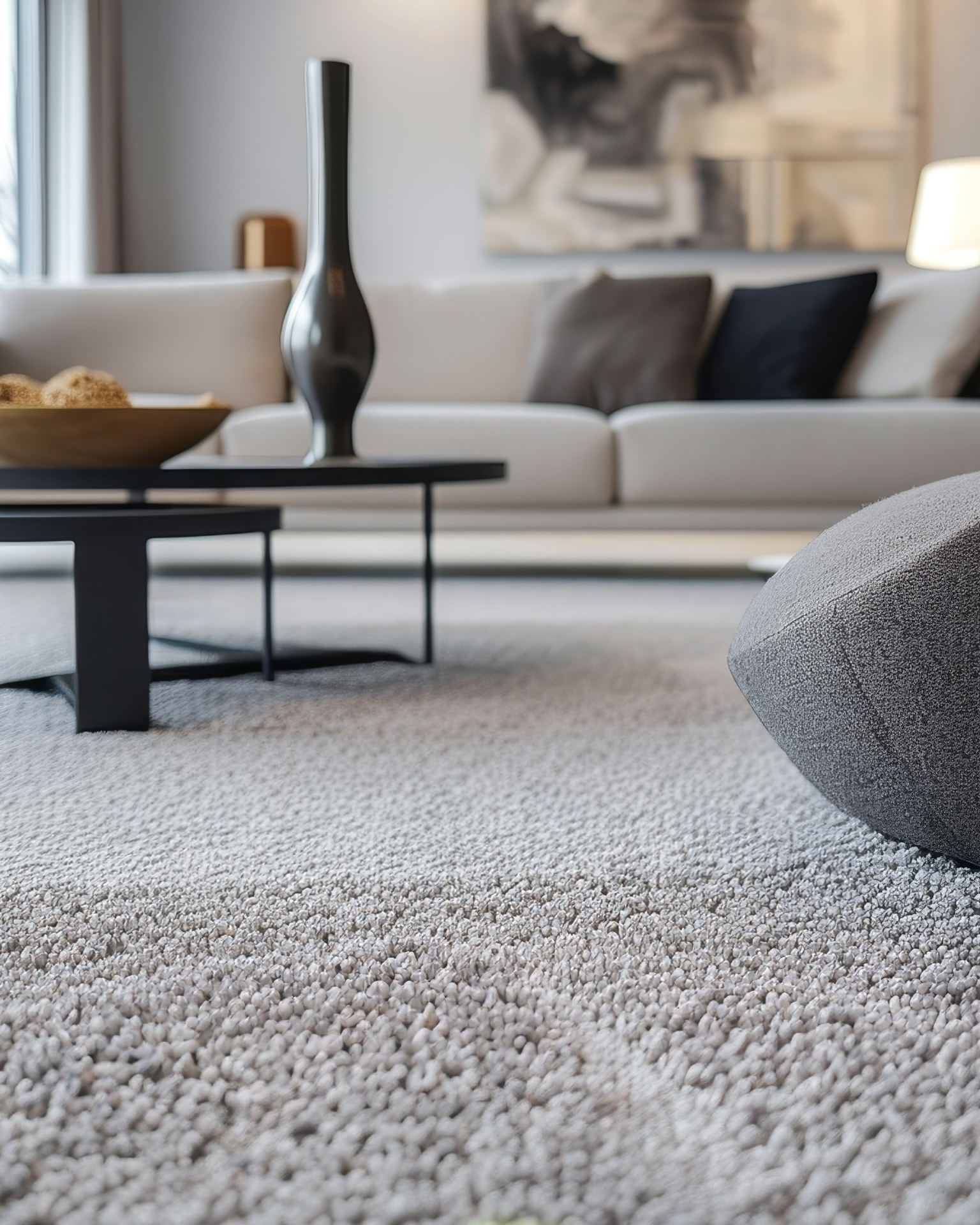 Close-up of a light grey textured carpet in a modern living room with a sofa, coffee table and decorative vase.